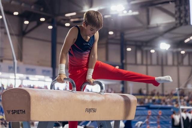 Young male gymnast performs a focused pommel horse routine, legs split wide, in a bright indoor sports hall.