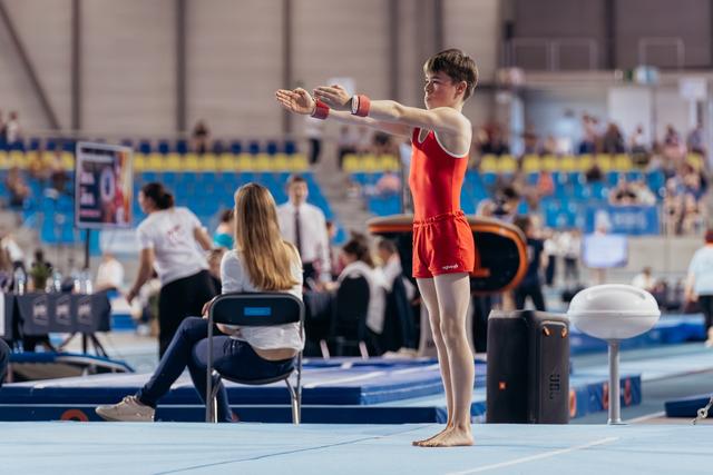 Young male gymnast in red leotard stands poised on floor mat, arms extended forward, focused expression before routine.