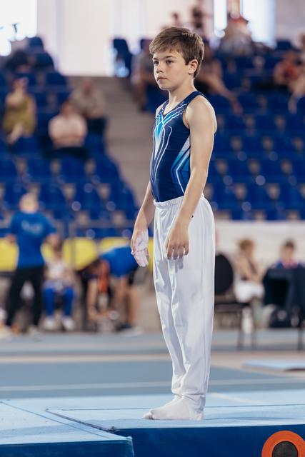 Young male gymnast stands still on the floor mat, focused and composed, wearing a blue leotard and white trousers.