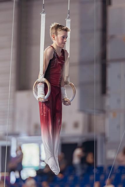 Young male gymnast in red uniform hangs still on rings, focused expression, competing in indoor gymnastics hall.