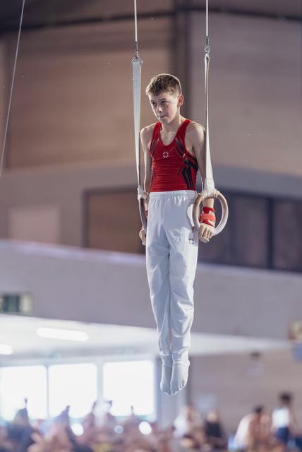 Focused young male gymnast in red leotard holds still rings with calm concentration in a competition gymnasium.