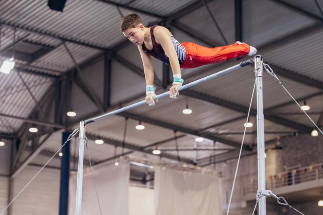 Young male gymnast performs on horizontal bar, body parallel, intense focus, inside a large indoor gymnasium.