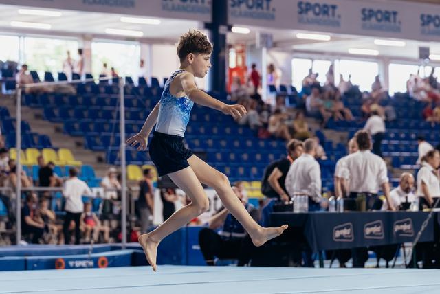 Young male gymnast leaps powerfully across the floor exercise mat, focused and composed at an indoor gymnastics meet.