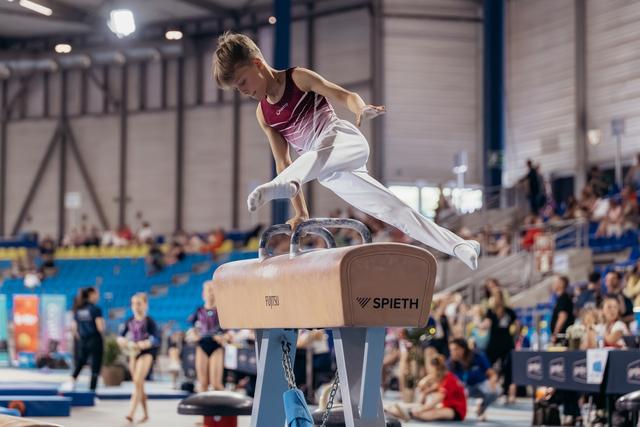 Young male gymnast performs on pommel horse, leaning forward with intense focus during an indoor gymnastics meet.