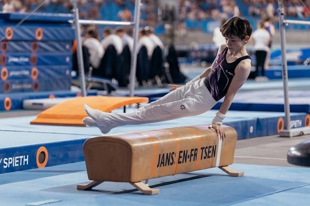 Young male gymnast performs a straddle hold on the pommel horse, legs extended, focused expression at an indoor competition.