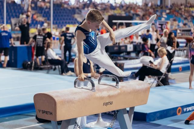Young male gymnast performs on pommel horse, leg raised high, intense concentration, in a busy competition arena.