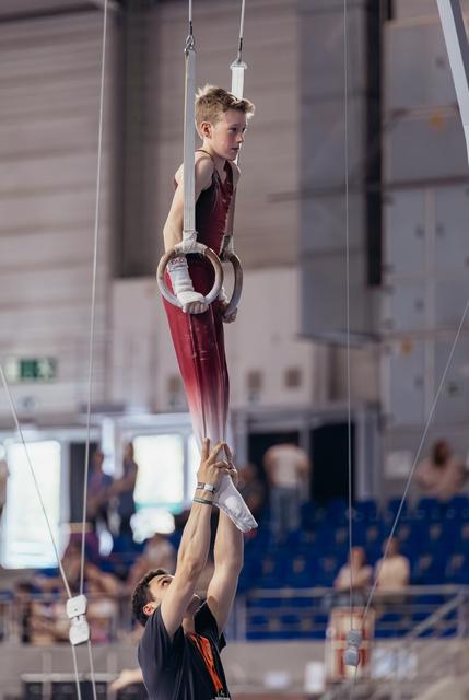 Young male gymnast hangs from still rings with intense focus while a coach supports his feet below in a sports hall.