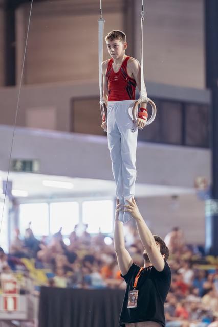 Young male gymnast holds still rings with intense focus while coach below steadies his feet in a packed arena.