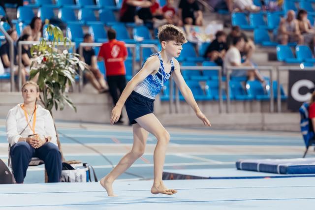 Young male gymnast runs barefoot across the floor exercise mat with focused concentration during a gymnastics meet.