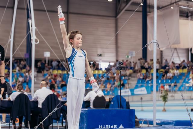 Young male gymnast raises hand to salute judges, standing confidently on podium in white and blue Agiva leotard.