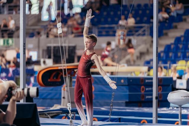 Young male gymnast raises one hand in a confident salute on the trampoline, wearing red suit and chalk-covered grips.