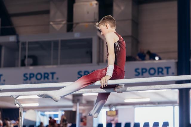 Young male gymnast in red leotard sits on parallel bars, looking down with focused concentration during a gymnastics meet.