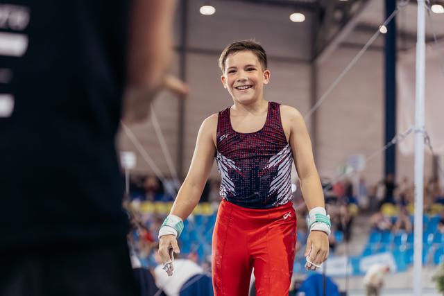 Young male gymnast beams with pride after his high bar routine, wearing red leo and wrist guards in a busy arena.