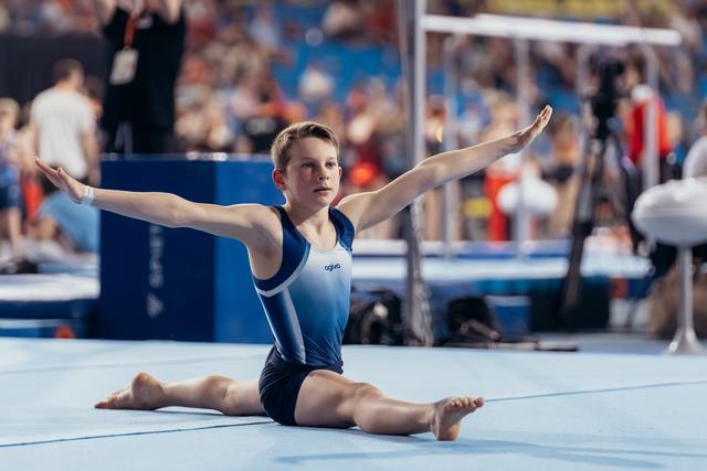 Young male gymnast performs a split on the floor with arms outstretched, displaying calm focus during a gymnastics event.