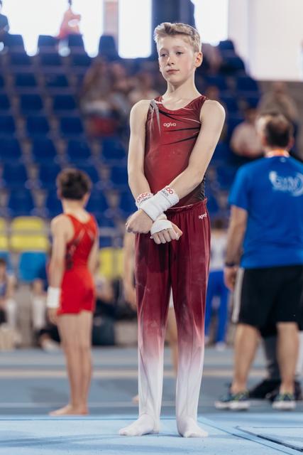 Young male gymnast in dark red agiva leotard stands composed, wrists wrapped, awaiting his turn on the competition floor.