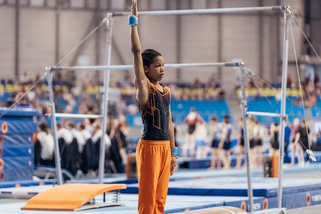Young gymnast stands focused beneath uneven bars, one arm raised, wearing orange pants and black leotard in busy gym hall.