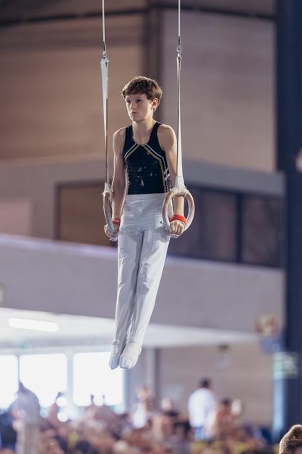 Young male gymnast holds still on rings, focused expression, wearing black leotard and white pants in a competition hall.