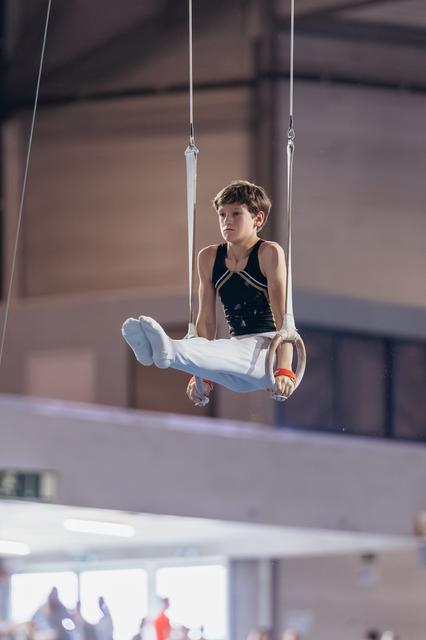 Young male gymnast holds a tucked seat position on still rings, focused expression, indoor gymnastics hall.