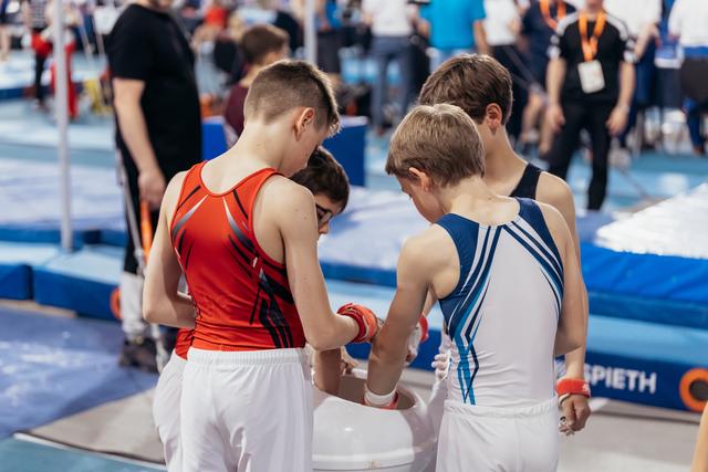 Young male gymnasts help each other wrap wrists before competing, showing camaraderie in a busy gymnastics hall.