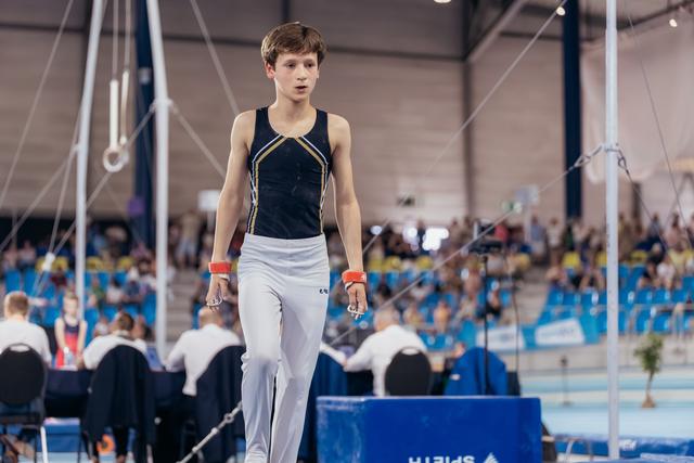 Young male gymnast walks with focused expression across the floor, wearing black leotard and white pants in a busy sports arena.