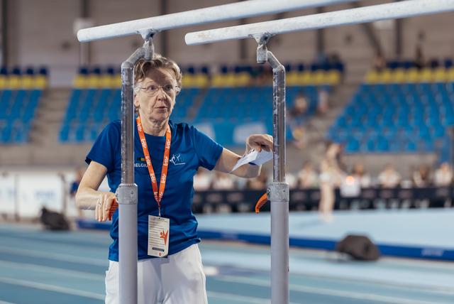 Older Belgian official stands focused between parallel bars in an indoor athletics stadium, holding a paper.
