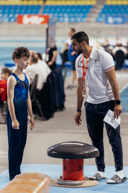 A young male gymnast listens attentively as his coach leans in to give instructions beside the vault apparatus.