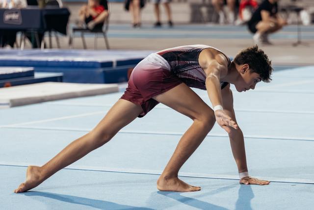 Young male gymnast barefoot on blue floor mat, crouched low in focused landing position during competition.