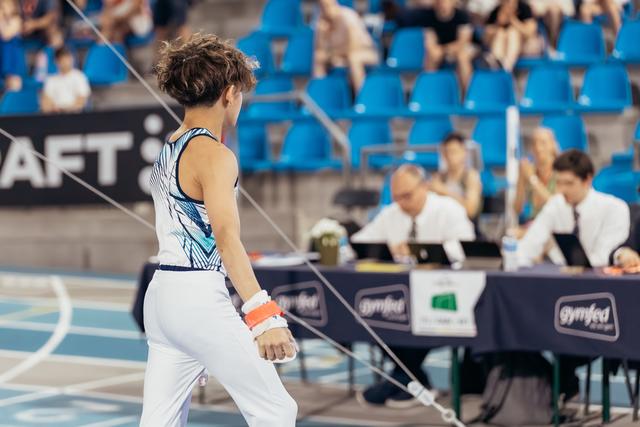 Young male gymnast stands with back to camera, facing judges at Gymfed table, composed before or after apparatus.