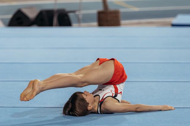 Gymnast performs a dramatic back arch on the floor exercise mat, legs raised high, face turned upward with focus.