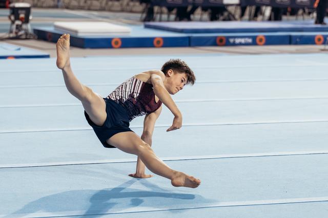 Young male gymnast mid-leap on floor exercise, body stretched low, intense focus on face during competition.