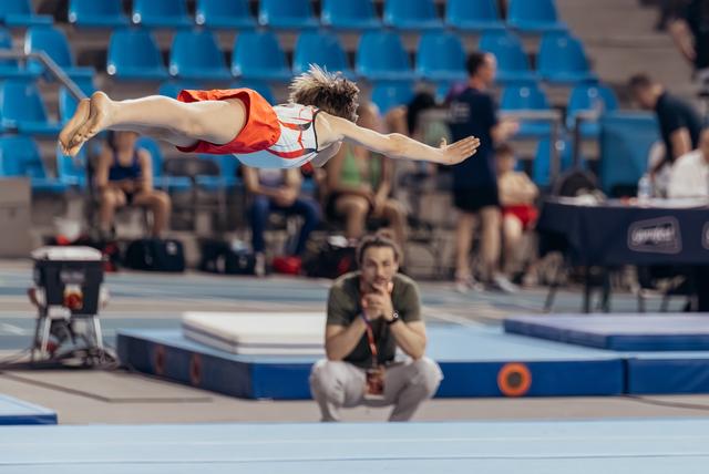 Young gymnast soars in a superman dive over the floor mat while a focused coach crouches and watches intently below.