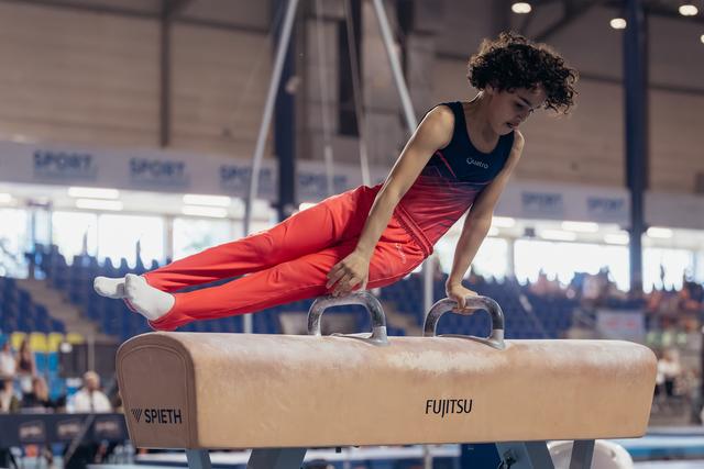 Young male gymnast with curly hair concentrates intensely while performing on the pommel horse at an indoor competition.