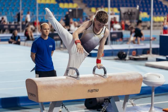 Young male gymnast performs scissors on pommel horse, concentrated expression, coach watching attentively in background.