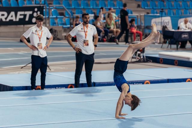 Young male gymnast performs a handstand on the floor while two coaches observe attentively in a blue competition arena.