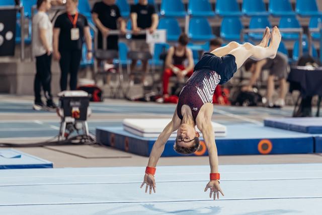 Young gymnast performing an inverted handstand on floor, wearing glasses and red wristbands, focused expression mid-routine.