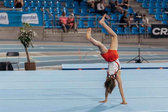 Young gymnast performs a one-handed cartwheel on the blue competition floor, legs split wide in a gymnastics arena.