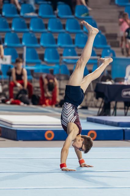 Young male gymnast performs a handstand with legs split on the floor exercise mat, wearing red wristbands.