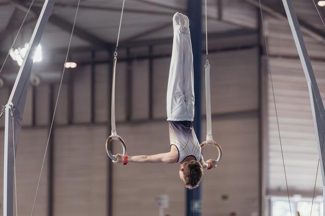Young male gymnast performs a perfect inverted hang on still rings, body fully extended, in an indoor gymnasium.