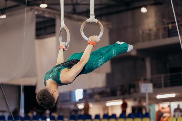 Young male gymnast holds an inverted position on still rings, body horizontal, wearing a teal leotard in a competition hall.
