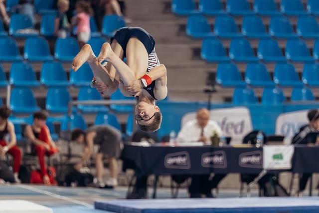 Young gymnast performs an inverted tucked flip mid-air, focused expression, blue arena seating in background.