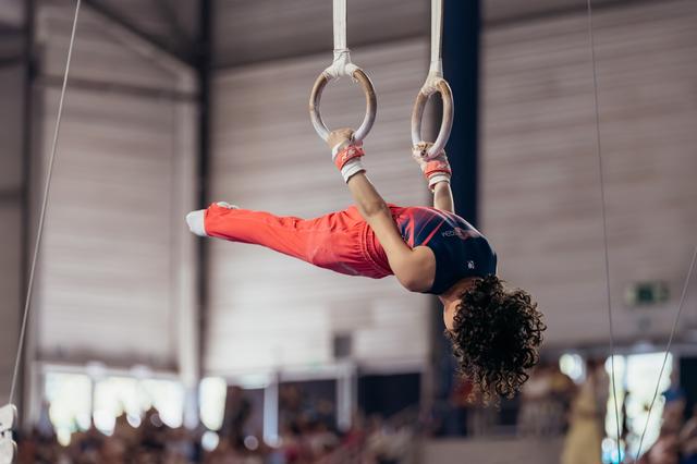 Young gymnast performs a dramatic iron cross hold on still rings, body fully inverted, showing strength and focus.