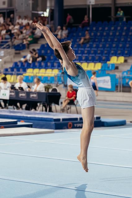 Young male gymnast mid-leap on floor exercise, arms reaching skyward, poised and focused during competition.
