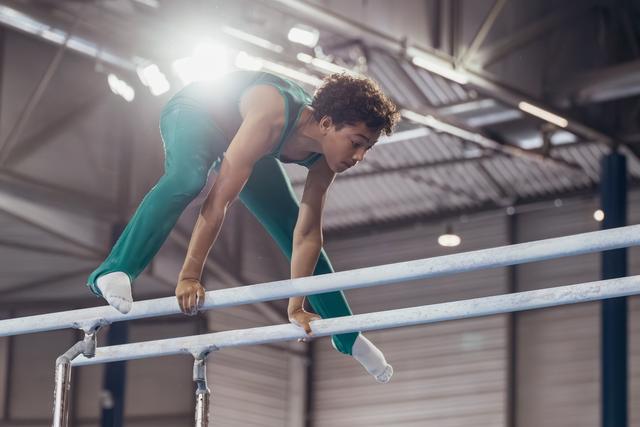 Young male gymnast in teal leotard leans forward on parallel bars with intense concentration in an indoor gymnasium.