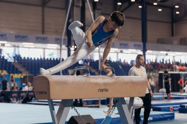 Young male gymnast performs on pommel horse, leaning forward with intense focus during an indoor competition.