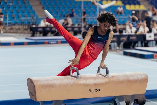 Young male gymnast performs a precise pommel horse routine, legs split wide, focused expression in a competition arena.