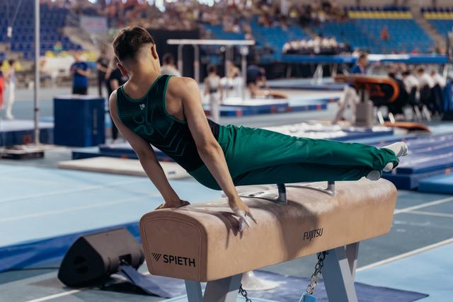 Young male gymnast in green leotard performs a straddle hold on the pommel horse during a competition.