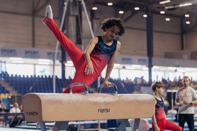 Young gymnast in red performs a focused routine on the pommel horse, one leg raised high in a competition hall.