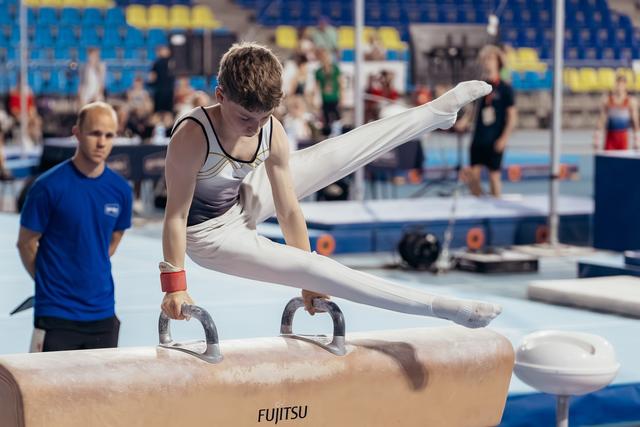 Young male gymnast performs a scissor split on the pommel horse, intense focus, while a coach watches nearby.