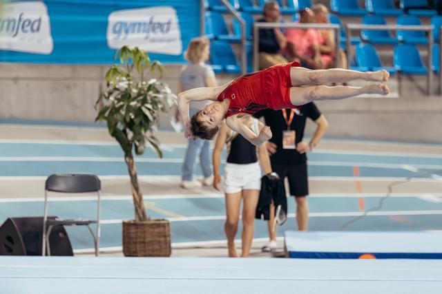 Young male gymnast in red leotard performs a graceful layout back flip mid-air at a Gymfed indoor competition.