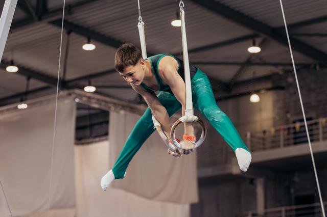 Young male gymnast grips still rings mid-routine, body coiled with intense focus, in a large indoor gymnasium.